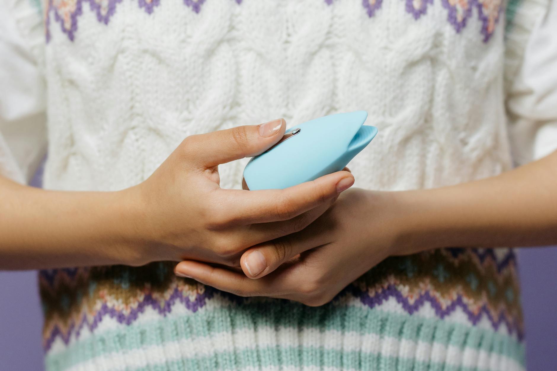 Hands holding a blue personal massager against a knitted sweater in intimate, private moment