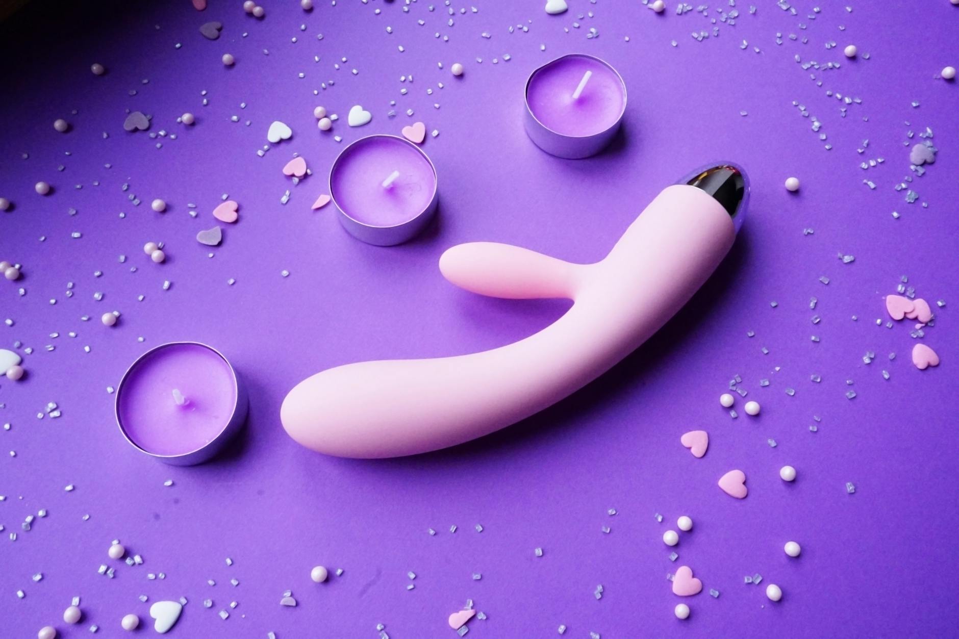 A pink vibrator on a purple background with romantic candles and heart confetti